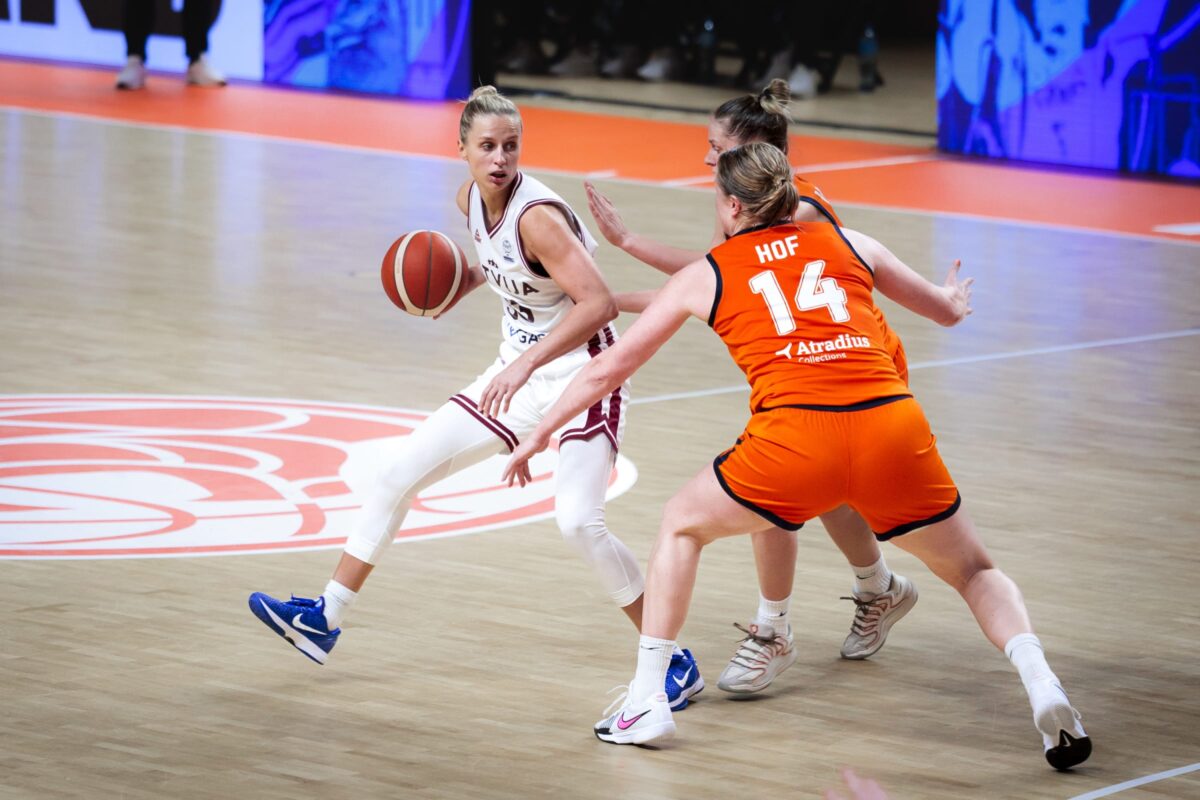 Latvijas basketbolistes cīnās par uzvaru Eiropas čempionāta kvalifikācijas spēlē uz basketbola laukuma fona.